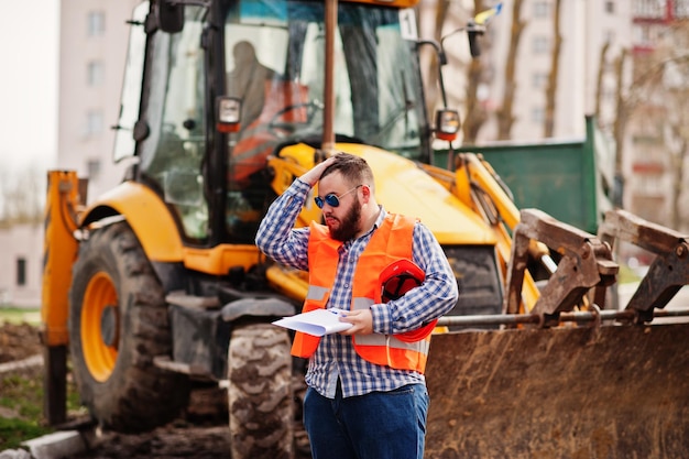 brutal-beard-worker-man-suit-construction-worker-safety-orange-helmet-sunglasses-against-traktor-with-plan-paper-hands_627829-11385